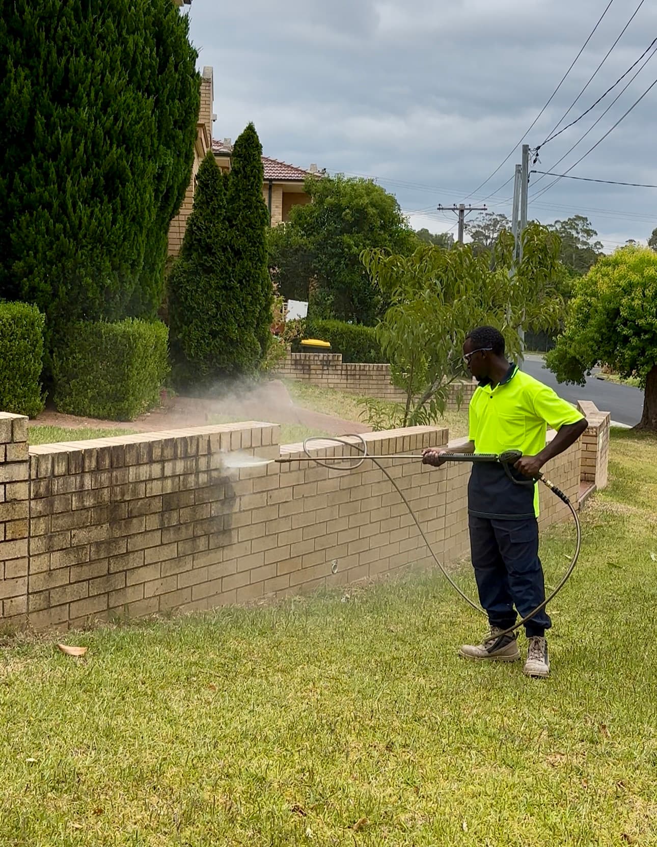 Worker in high-visibility shirt pressure washing a stained brick wall in a suburban yard.