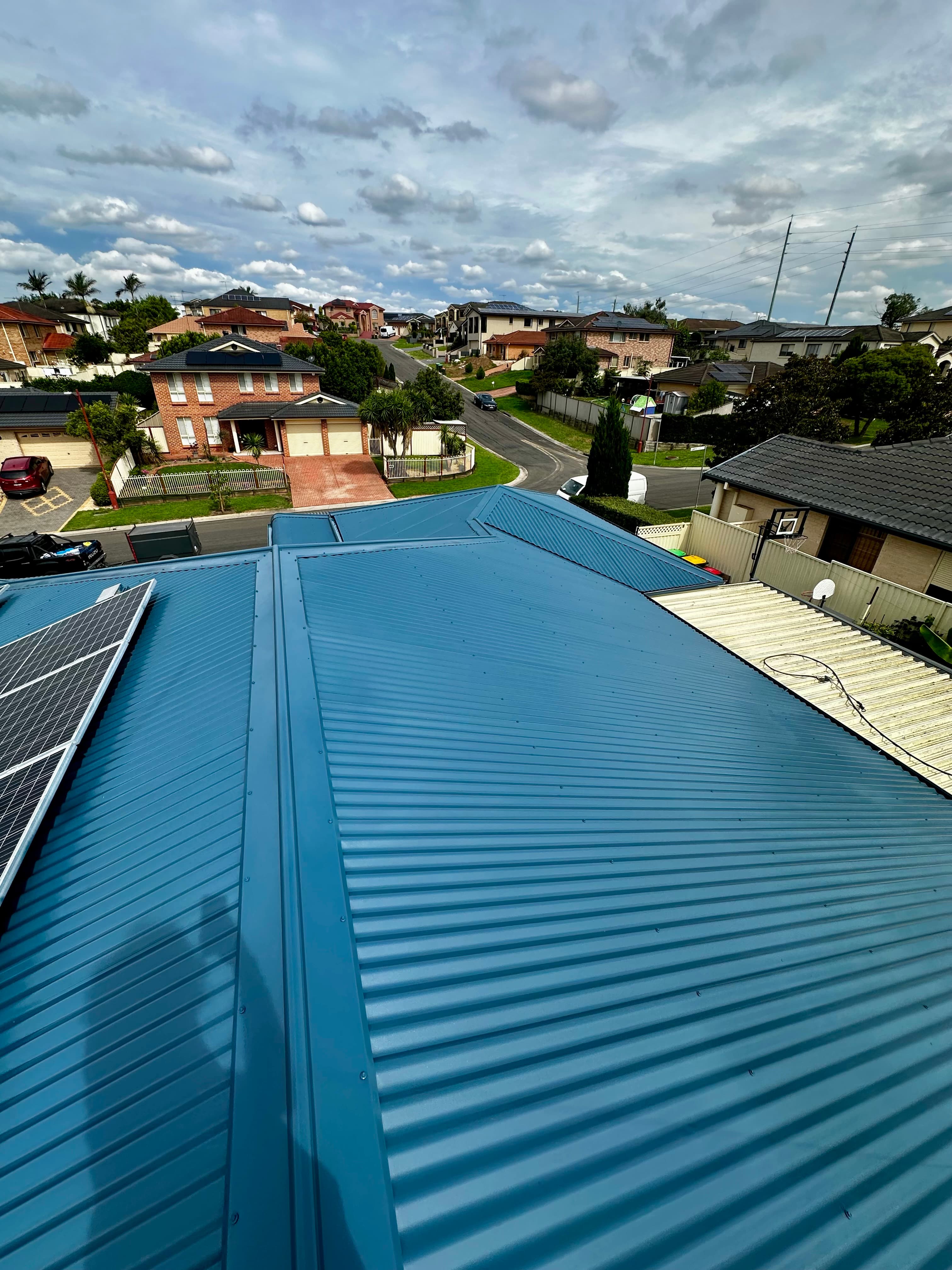 Blue corrugated metal roof with solar panels overlooking a suburban neighborhood under a cloudy sky.