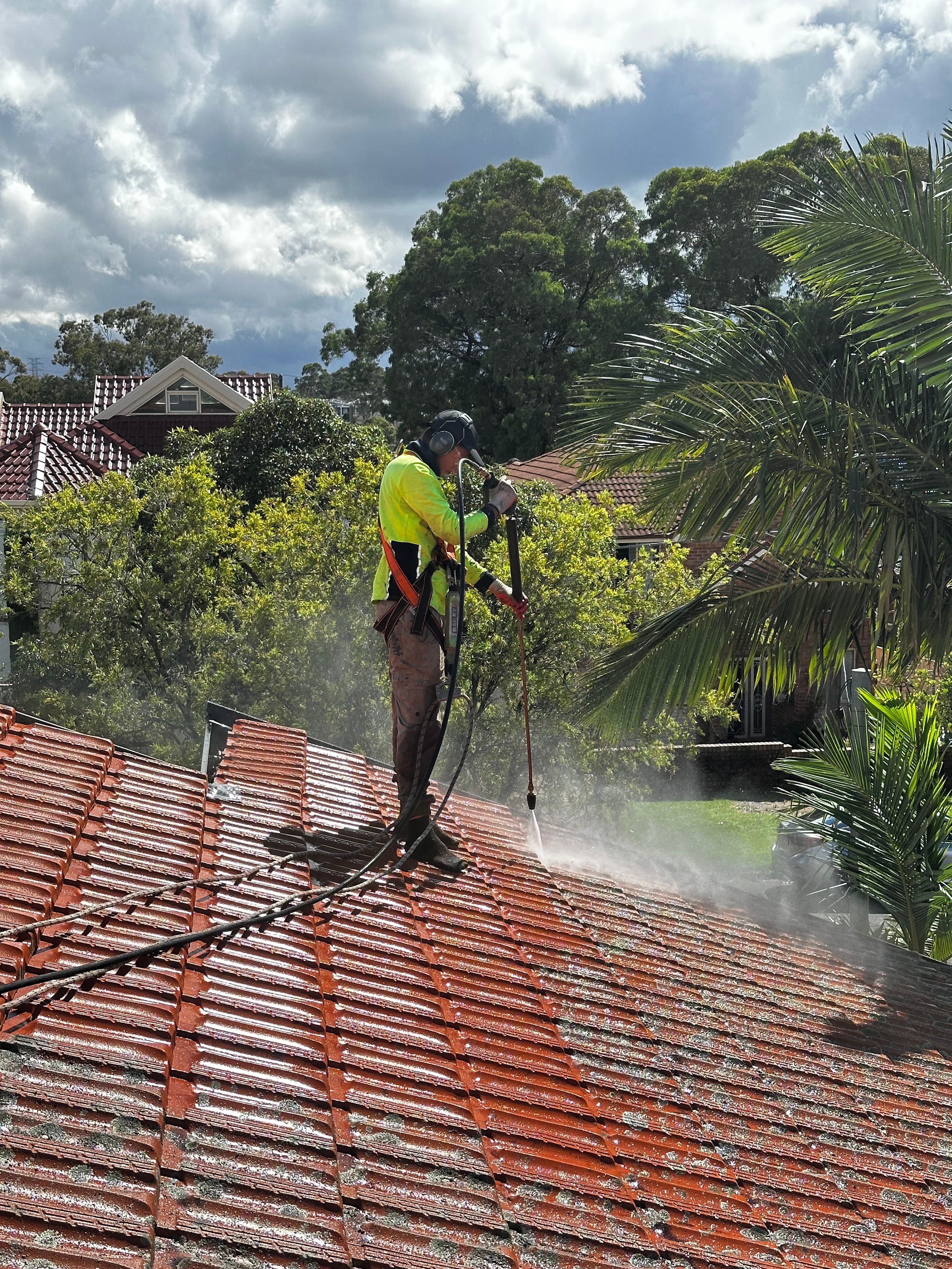 Worker in high-visibility gear pressure washing red terracotta roof tiles on a residential house.