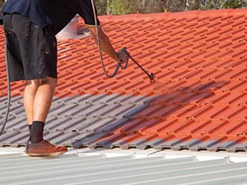 A worker applies bright orange paint to grey roof tiles using a spray gun.